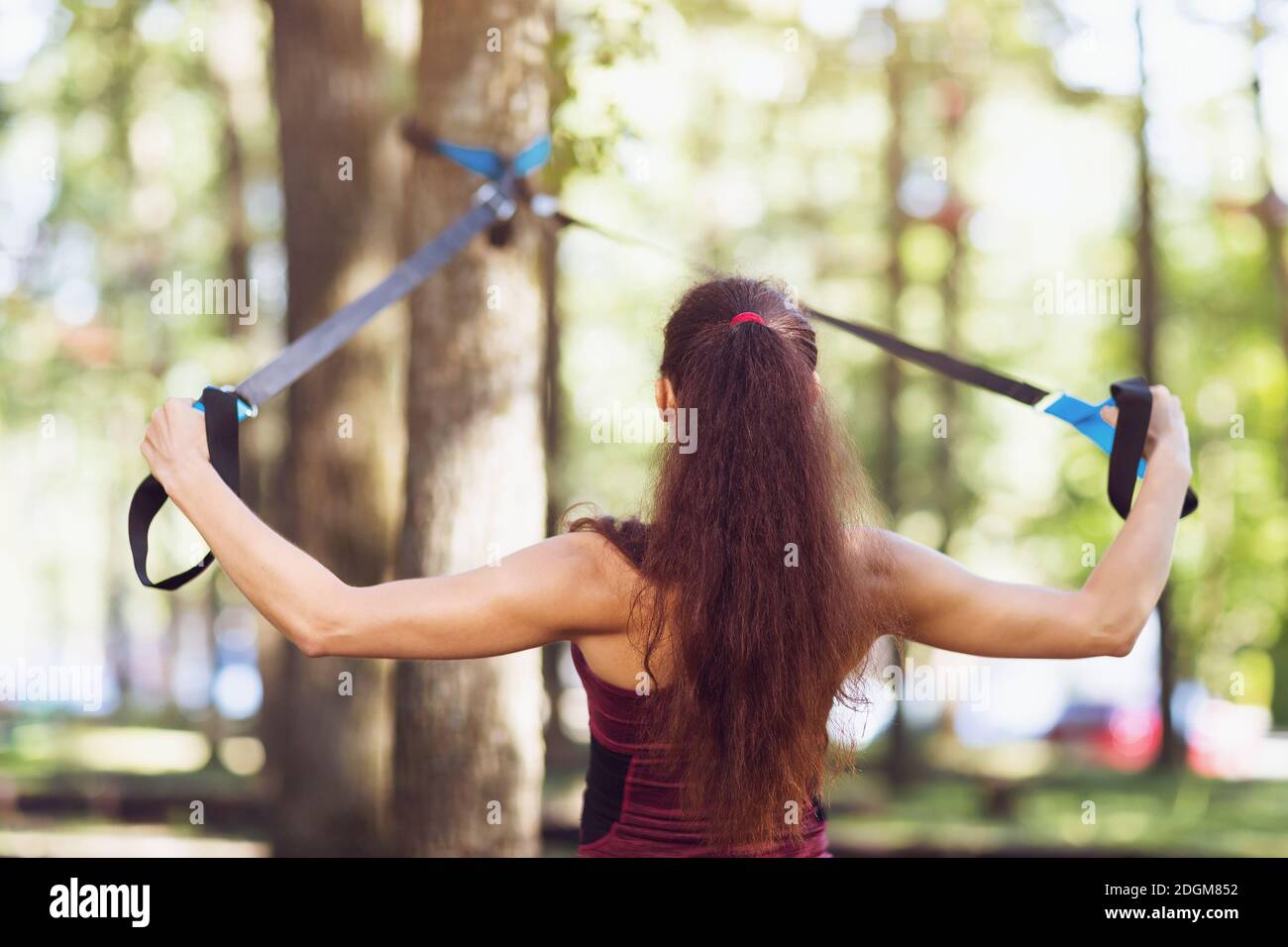 A young woman performs an exercise to work out the muscles of the back on an overhead machine attached to a tree in the park. Stock Photo