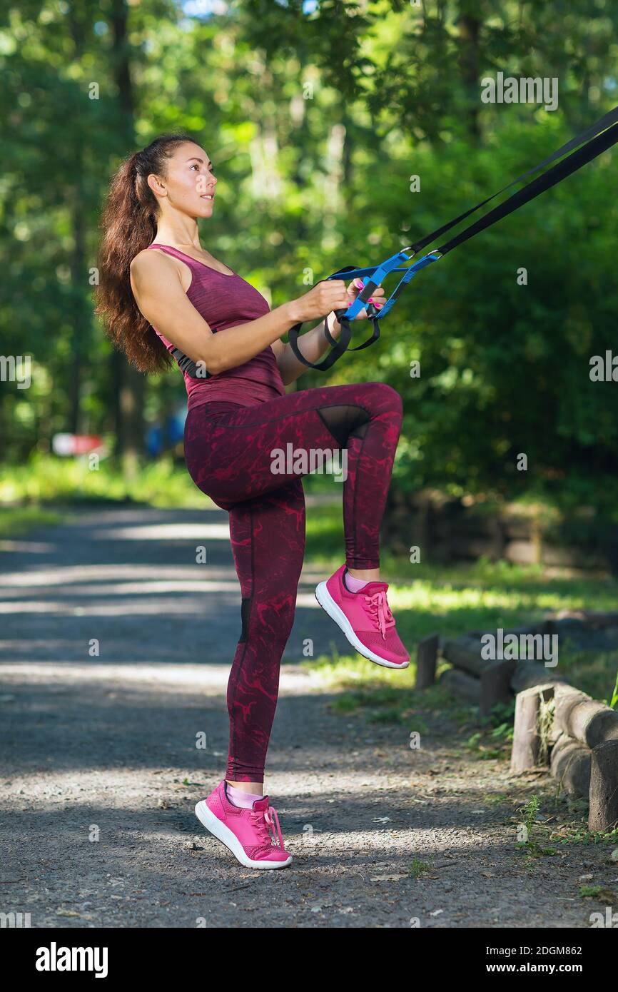 A young woman performs a muscle training exercise on a suspended machine attached to a tree in a park. Stock Photo