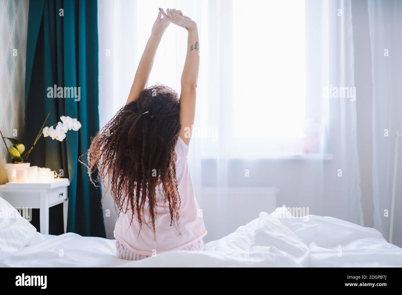 Young long-haired woman outstretching her arms sitting on the bed after good night sleep in light modern bedroom, back view Stock Photo