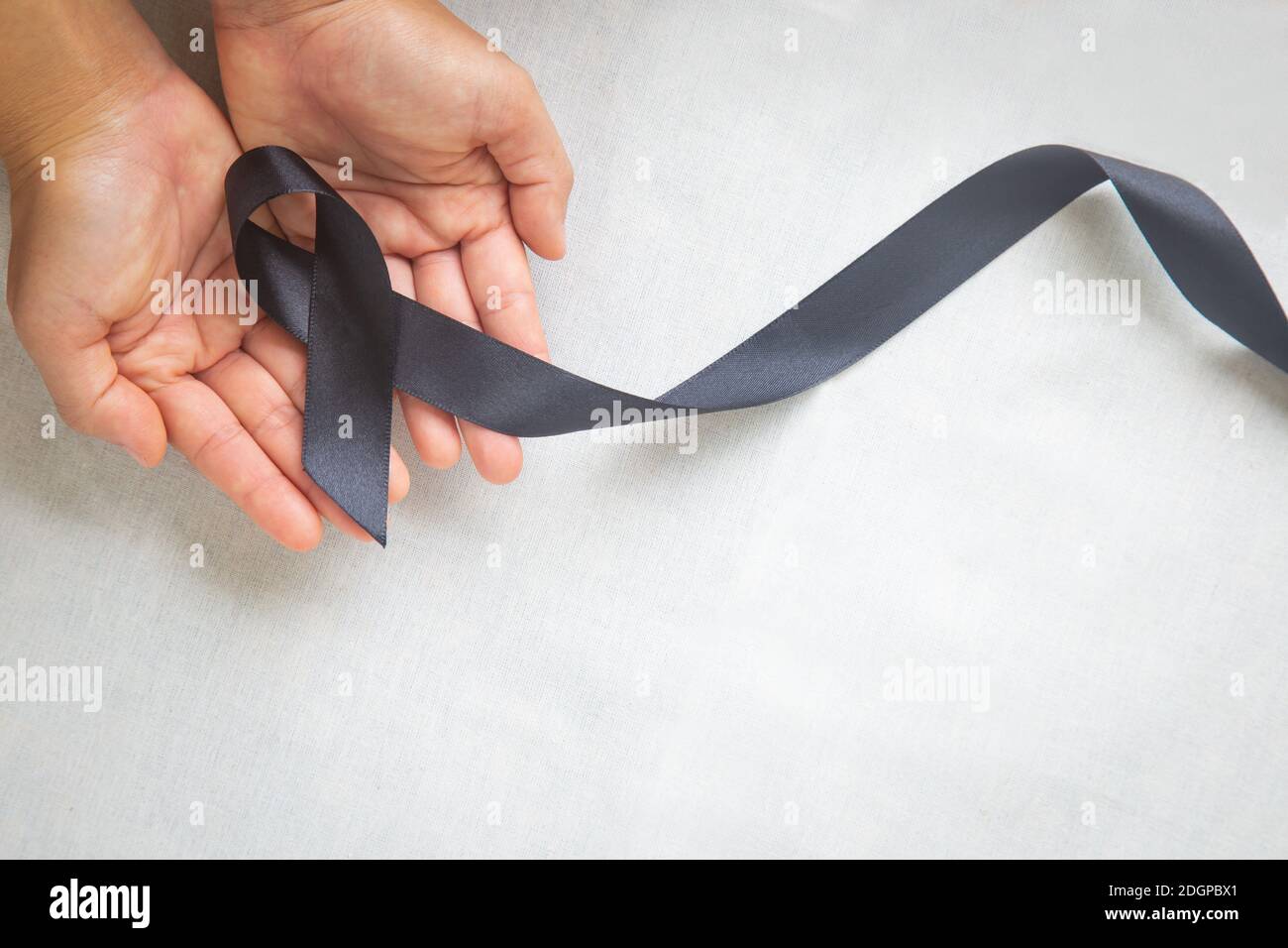 Hand holding Black ribbon on white fabric background with copy space, symbol of Skin Cancer awareness month on May, Melanoma cancer, Mourning ribbon s Stock Photo