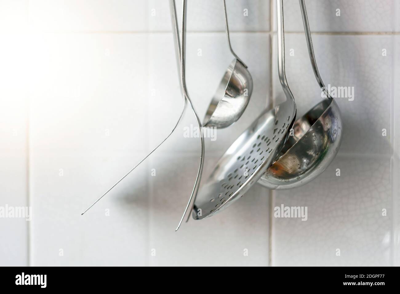 A skimmer, two forks and two metal ladles hanging on the tiled kitchen wall. Metal kitchen utensils Stock Photo