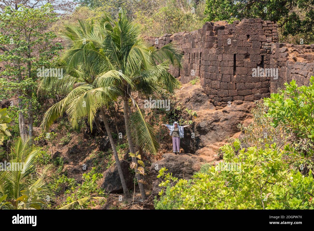 A woman on a rock next to an old stone tower and the walls of the Portuguese fortress Fort Cabo De Rama in the rainforest in Goa, India Stock Photo
