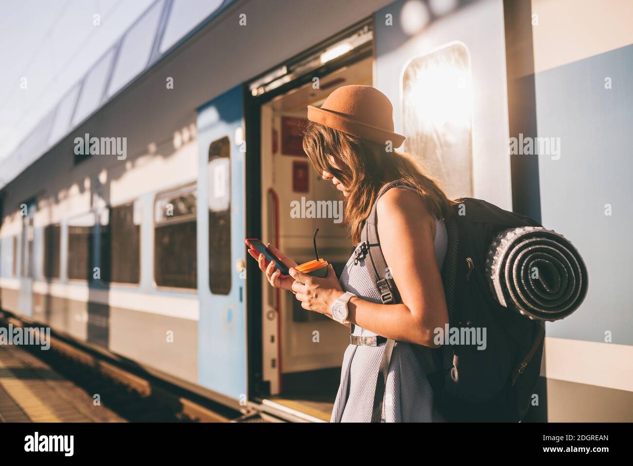 Railroad theme. Beautiful young woman with a backpack uses the phone while standing near the railroad train on the platform. Che Stock Photo