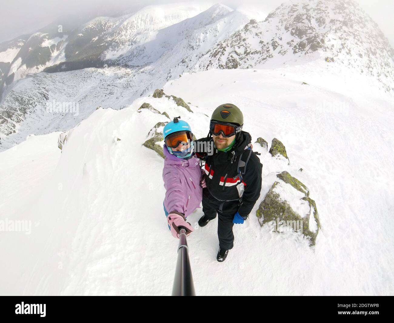 Young and happy guy and girl make a photo with a self-stick stick and smartphone on the peak of a snow capped mountain in a ski Stock Photo