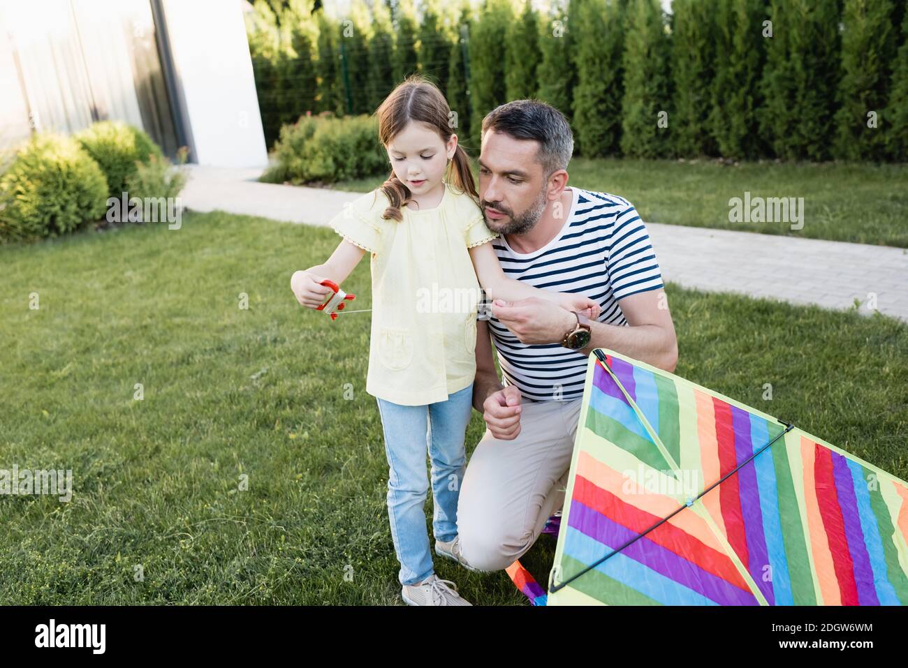 Daughter with kite string holder standing with father squatting near on lawn on blurred background Stock Photo