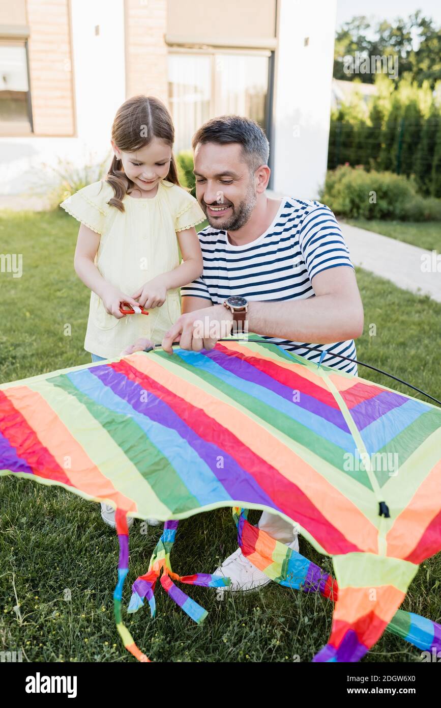 Full length of smiling daughter standing near father assembling kite on lawn with blurred house on background Stock Photo