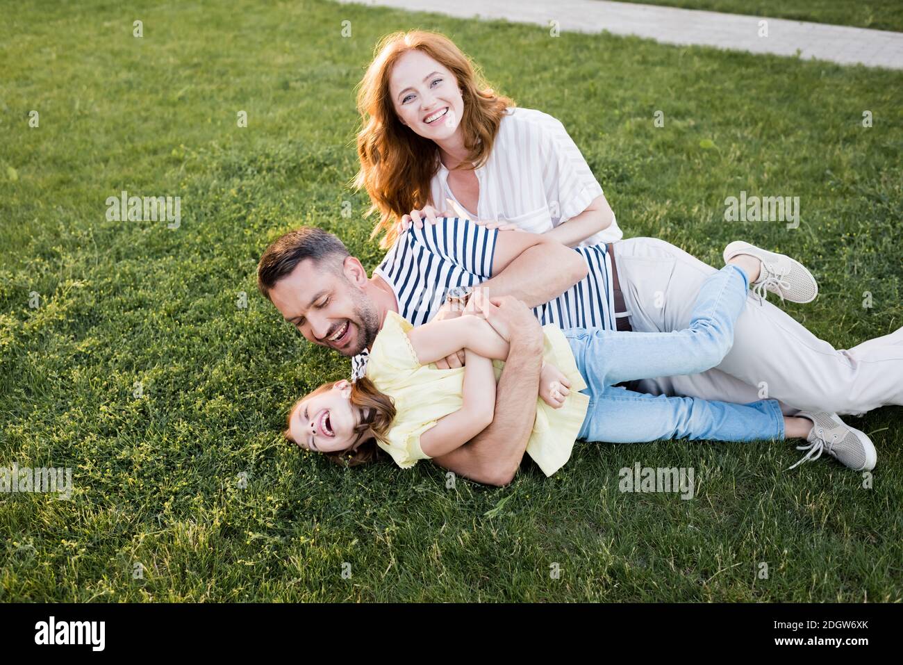 Happy redhead woman looking at camera near man tickling girl while lying on  lawn Stock Photo - Alamy