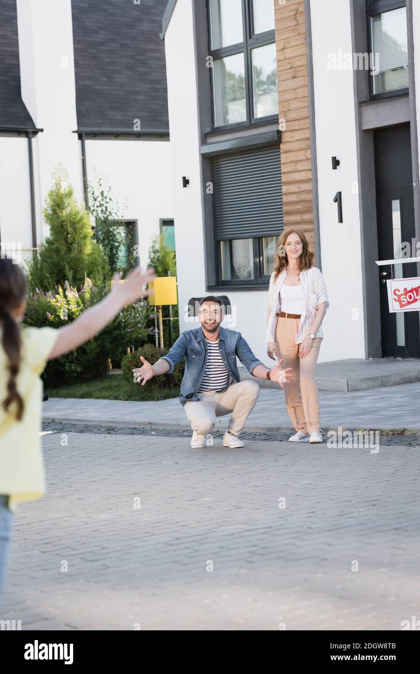 Redhead woman standing near man squatting with open arms while looking at daughter on blurred foreground Stock Photo