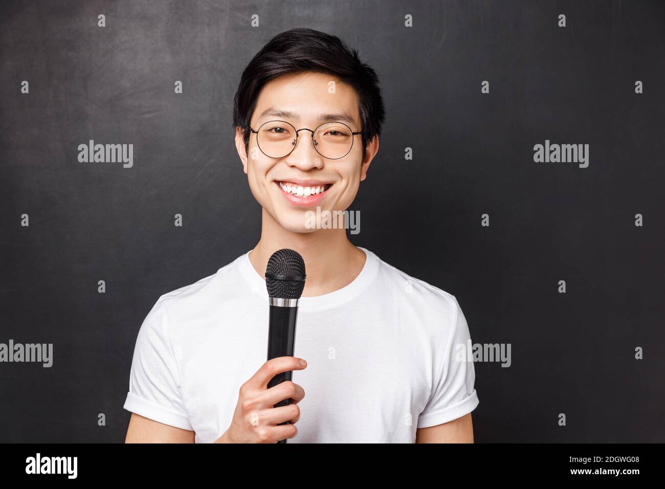 Leisure, people and music concept. Handsome and cute smiling asian man in white t-shirt, glasses, holding microphone, singing at Stock Photo