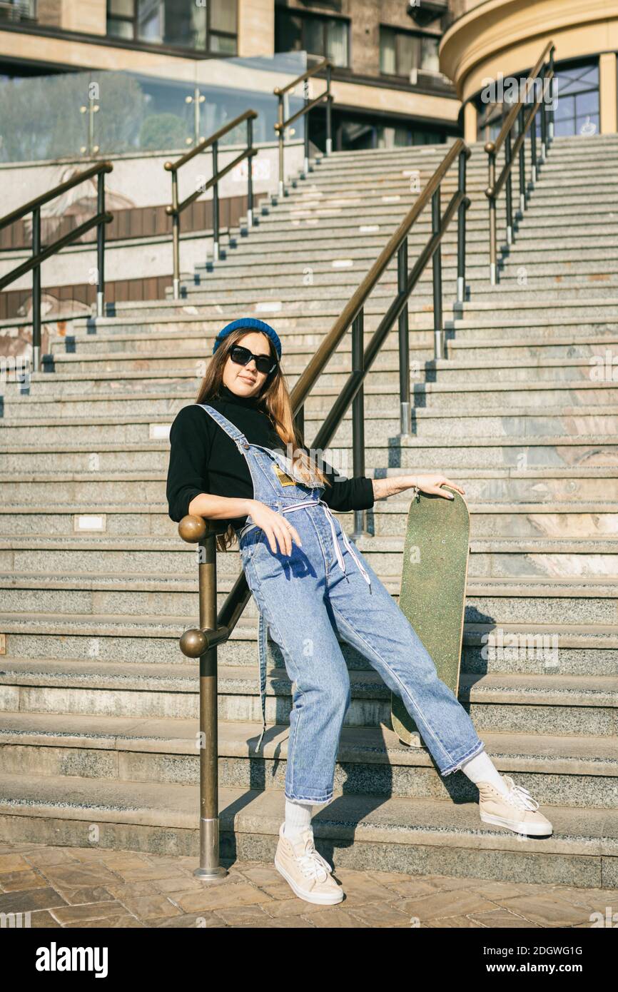 Stylishly dressed woman in blue denim jumpsuit posing with skateboard.  Street photo. Portrait of girl holding skateboard. Lifestyle, youth concept  Stock Photo - Alamy