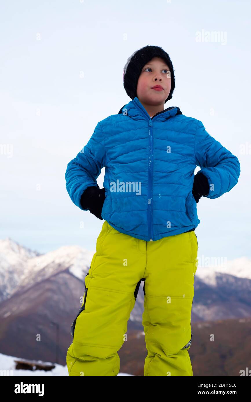 A boy in a blue jacket, yellow ski pants and a knitted hat stands against the backdrop of snow-capped mountains. Blue eyes look into the distance. Stock Photo