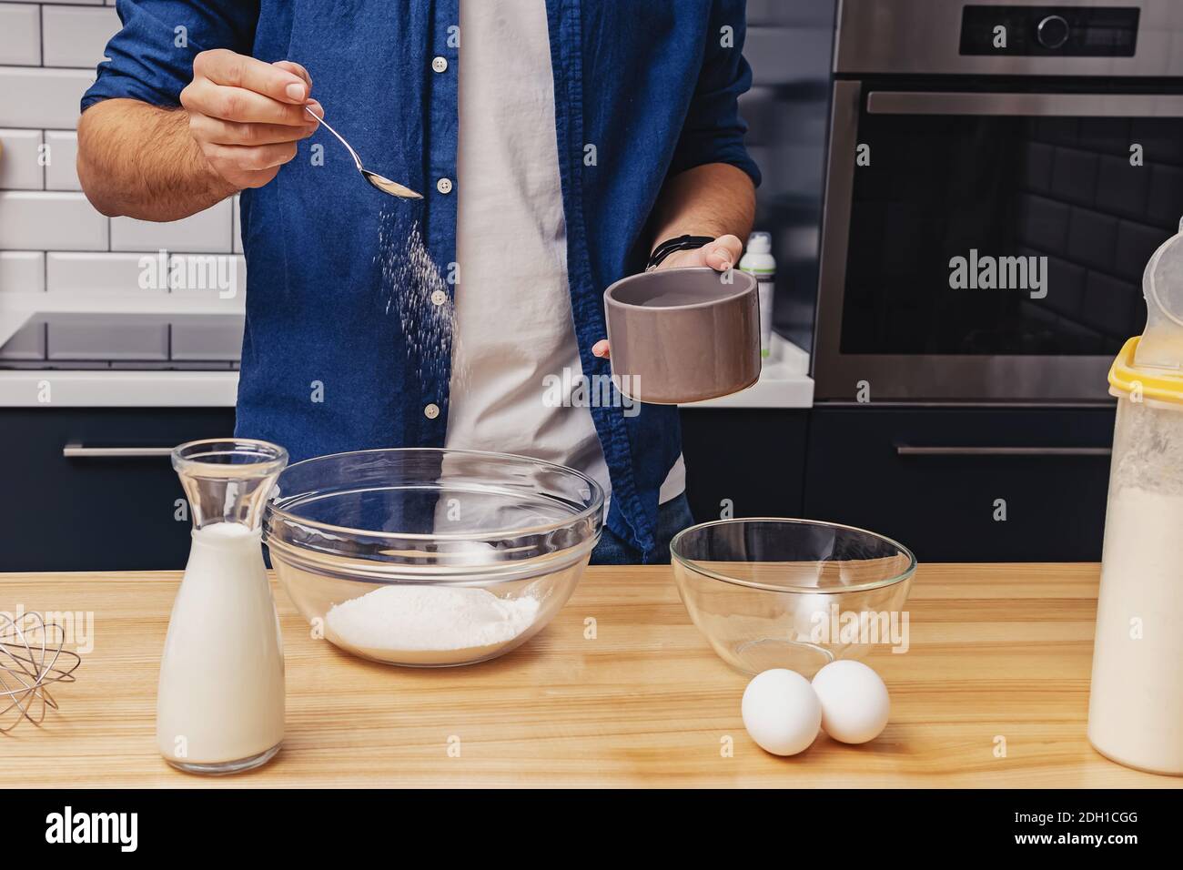 Young man cooking at his kitchen. Making pancakes or other dessert, adding sugar to bowl Stock Photo
