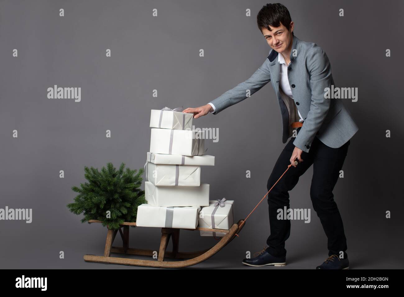 Christmas themed horizontal business portrait of a smart casual dressed female executive pulling a sleigh loaded with white and silver wrapped gift boxes on a grey background. Stock Photo