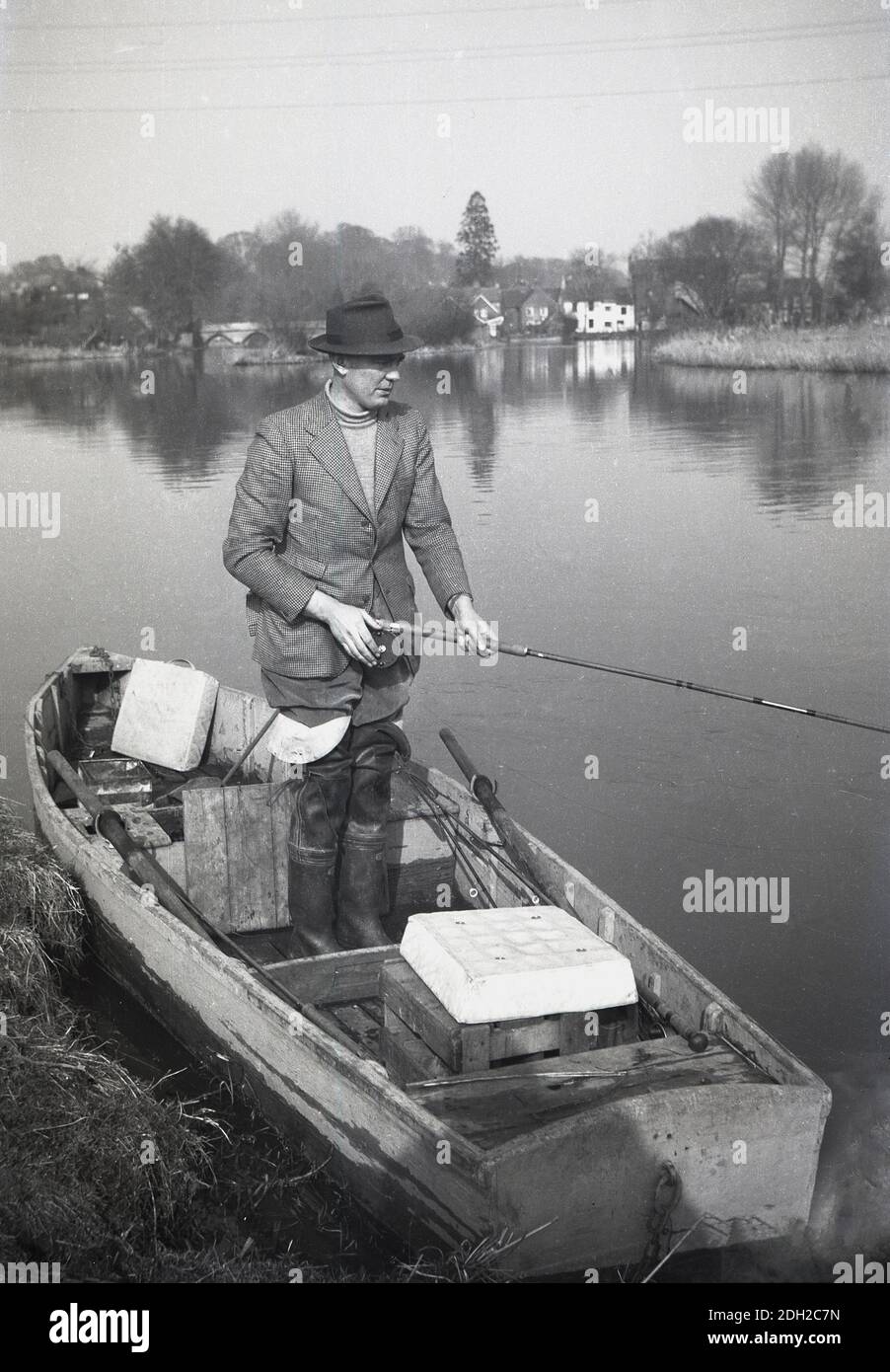 1950s, historical, on a calm English river, a picture of a gentleman fishing. Standing in a small wooden boat by the riverbank, and wearing long knee length wellington boots, a check jacket and hat, with fishing rod in hat, England, UK. Stock Photo