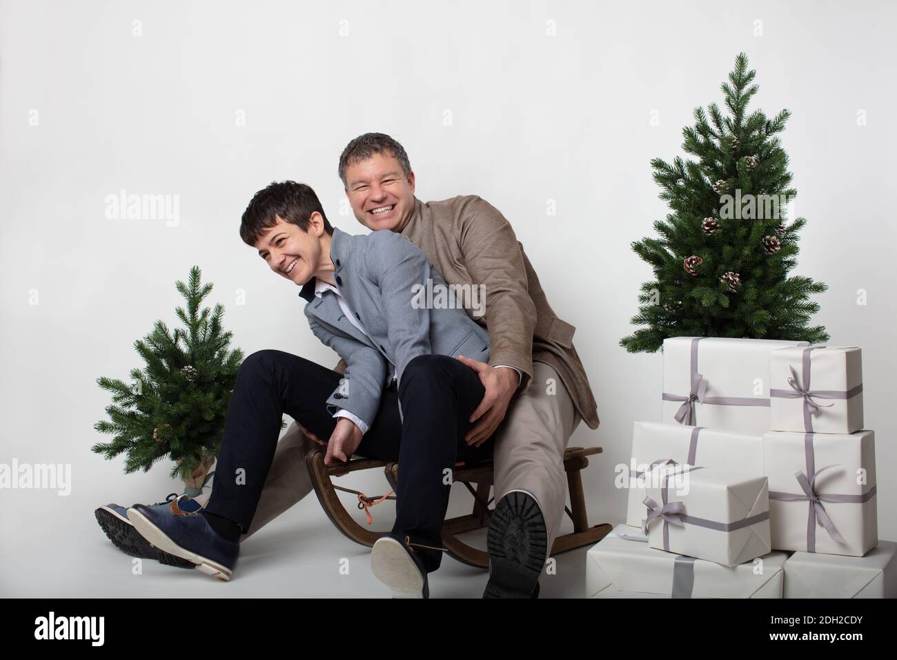 Christmas themed horizontal business couple portrait dressed smart casual in blue, white, beige pants and blazers on grey background. In the foreground a wooden sleigh with a pile of white gift boxes on it. Two miniature Christmas pine trees. Stock Photo