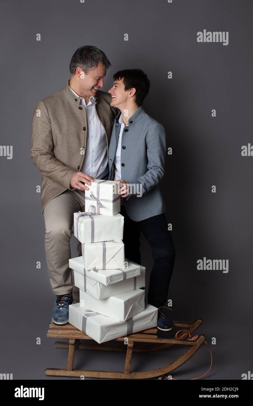 Christmas themed vertical business couple portrait dressed smart casual in blue, white, beige pants and blazers on grey background. In the foreground a wooden sleigh with a pile of white gift boxes on it. Stock Photo