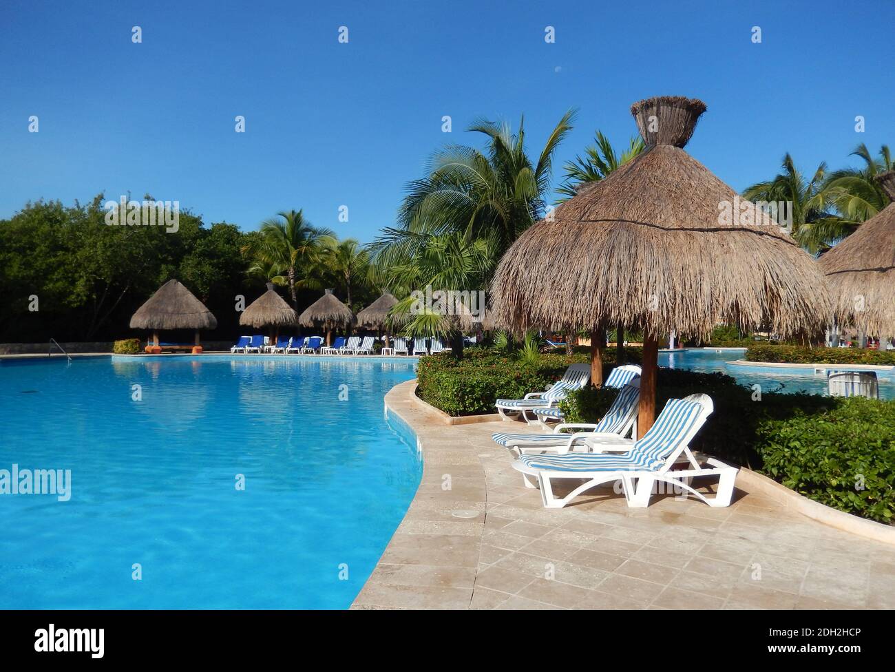 Pool sitting area with straw umbrellas, chaise longue chairs and palm trees landscape, at a Caribbean beach resort on the Riviera Maya, in Cancun, Mex Stock Photo