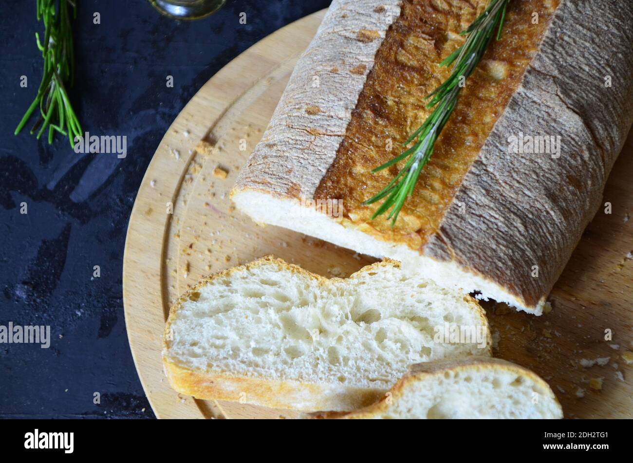 Italian ciabatta bread cut in slices on wooden chopping board with herbs, rosemary garlic and olives over dark grunge backdrop, top view. Stock Photo