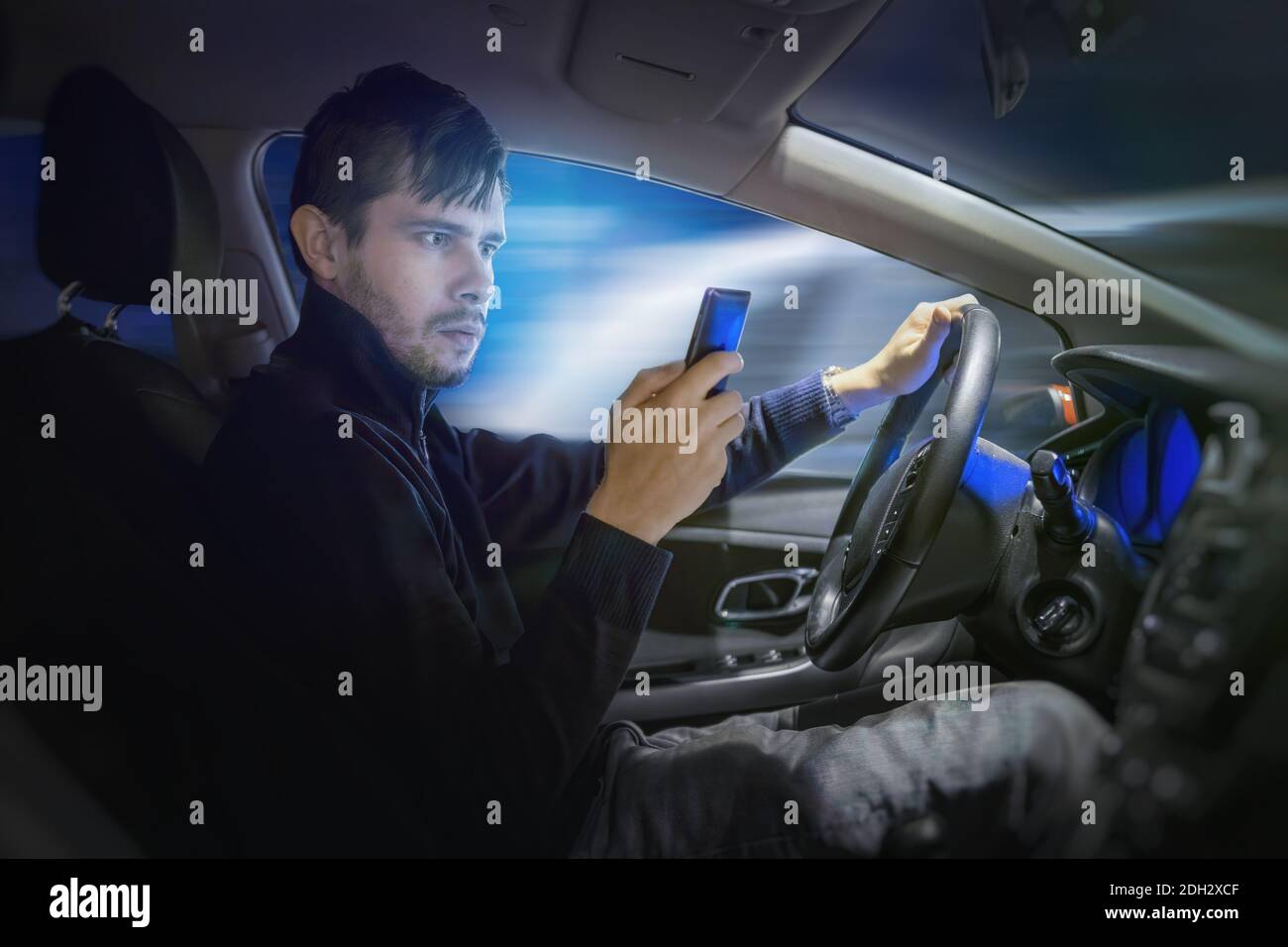 Young man is texting using smartphone and driving a car at night. Stock Photo