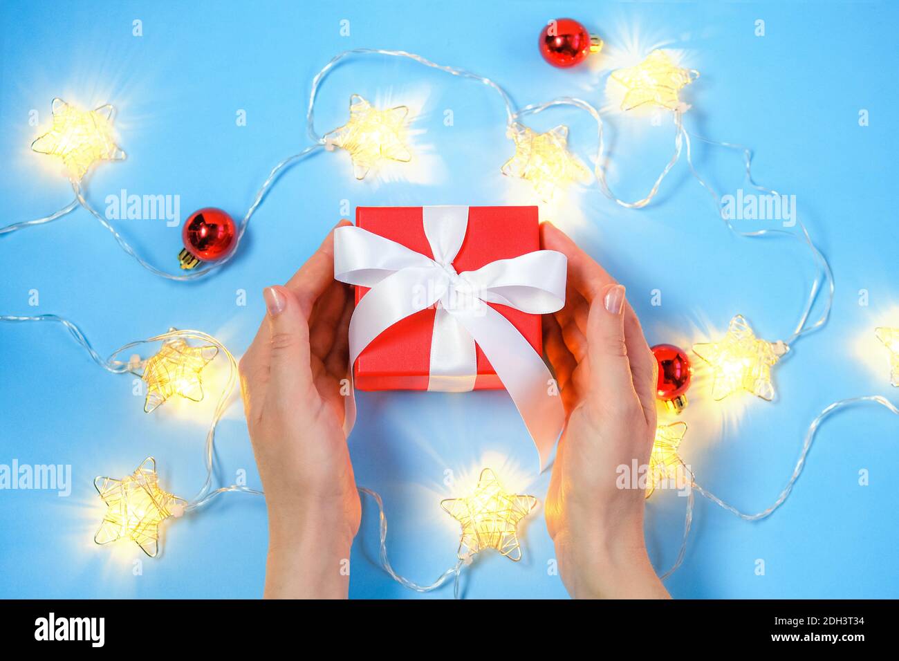 Woman's hands taking or giving colorful red present with luxury white ribbons on lignt blue background with Christmas decoration. Stock Photo