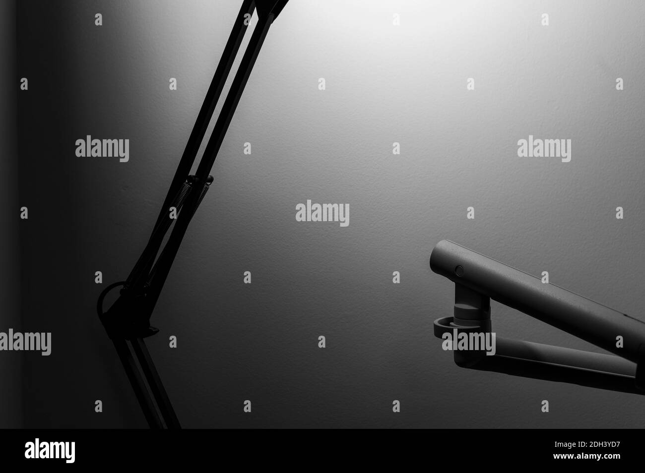 Black and white monochrome image of articulated desk lamp with black arms in foreground blur and with light bulb illuminated with its shadow on wall. Stock Photo