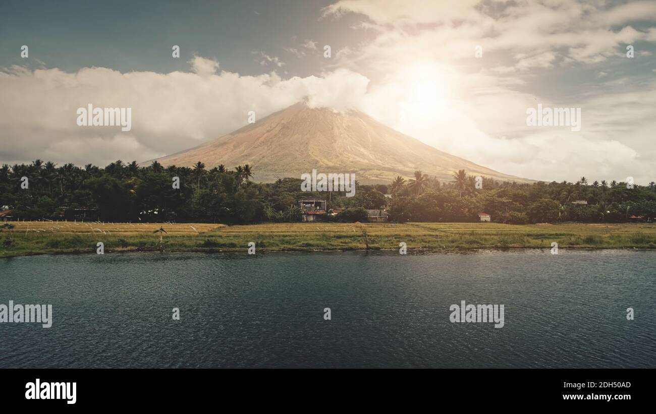 Volcano erupt at sun shine over lake shore aerial. Philippine coutryside of Legazpi town at green valley with palm trees. Rural cottages at exotic forest. Cinematic nobody nature landscape at sunlight Stock Photo