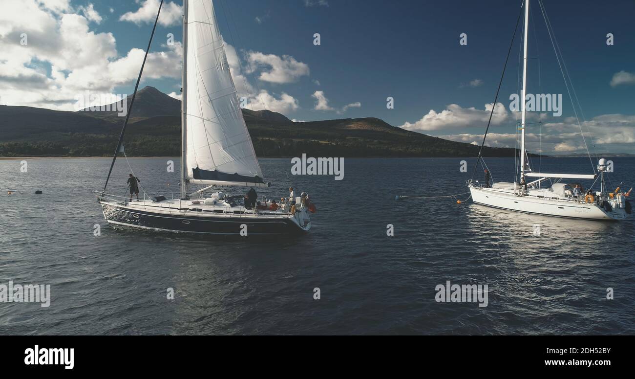 Closeup aerial of yachts cruising at ocean bay coast of mountain island of Arran, Scotland. Amazing seascape with passengers on sail boat. Cinematic ships race at open sea at drone shot Stock Photo