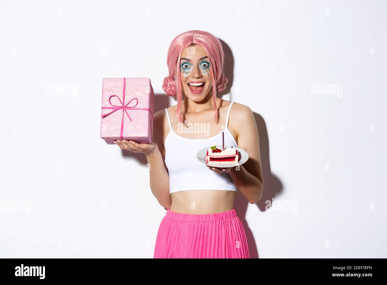 Portrait of surprised beautiful girl in pink wig, receive birthday gift, holding b-day cake and smiling happy, standing over whi Stock Photo