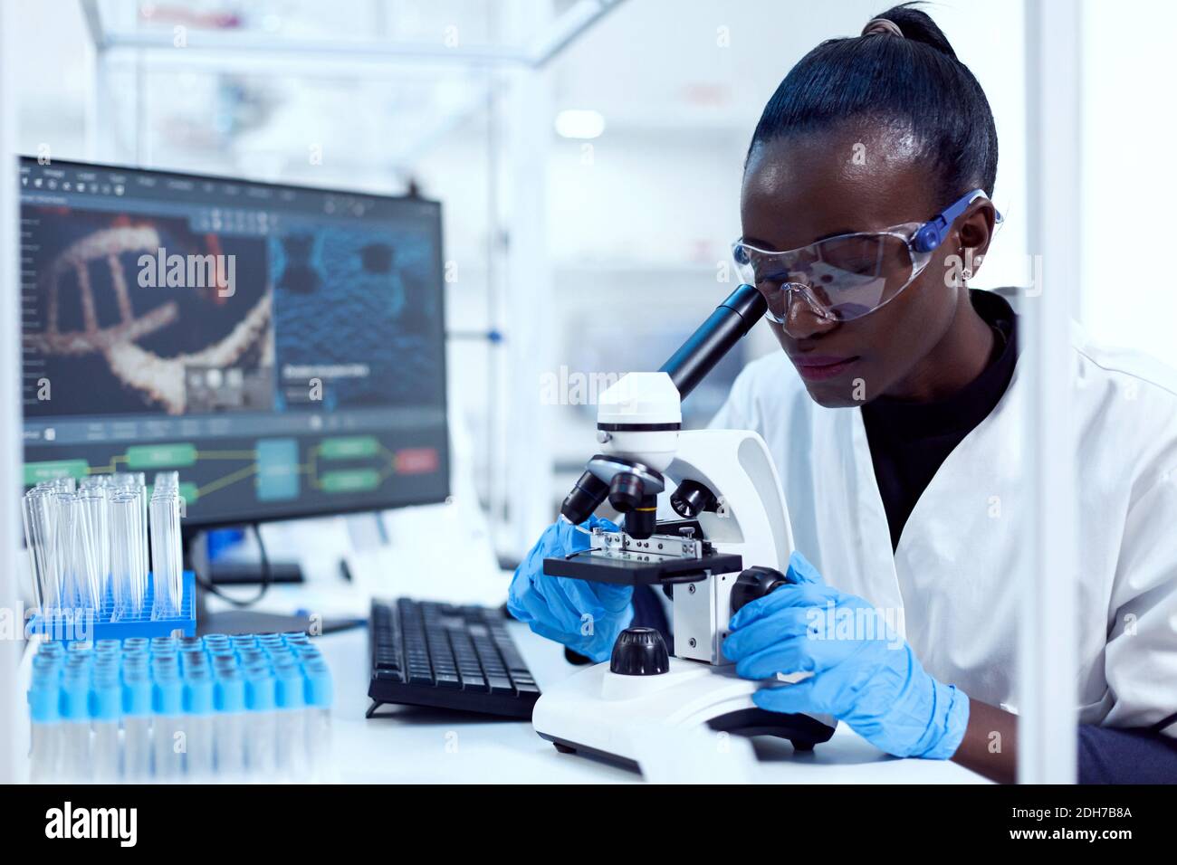 Virologist of african ethnicity sitting at her workplace neat test tubes looking through microscope. Black healthcare scientist in biochemistry laboratory wearing sterile equipment. Stock Photo