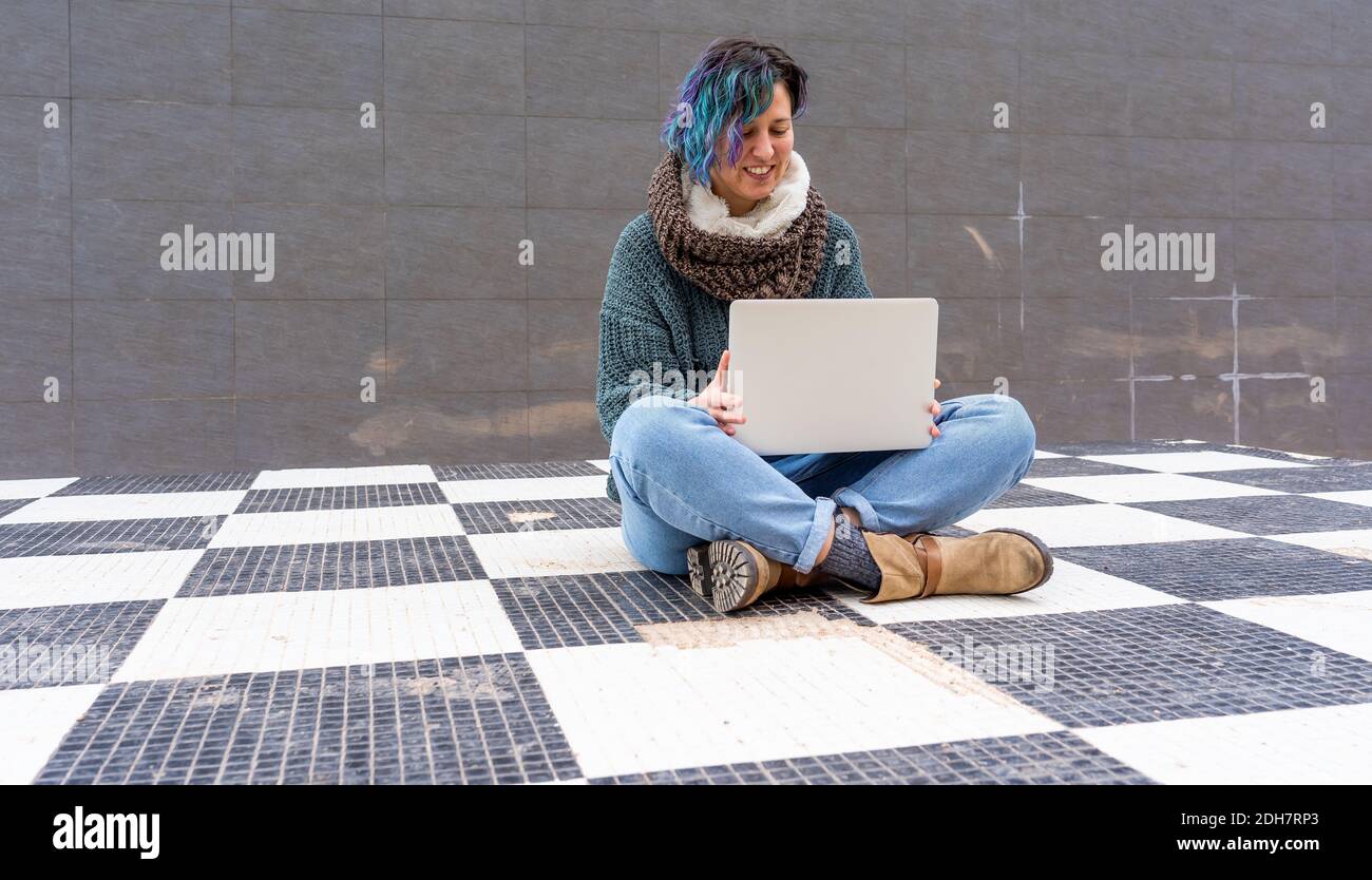 A stylish young lady with colorful hair sitting on a checkered floor in a park using her laptop Stock Photo