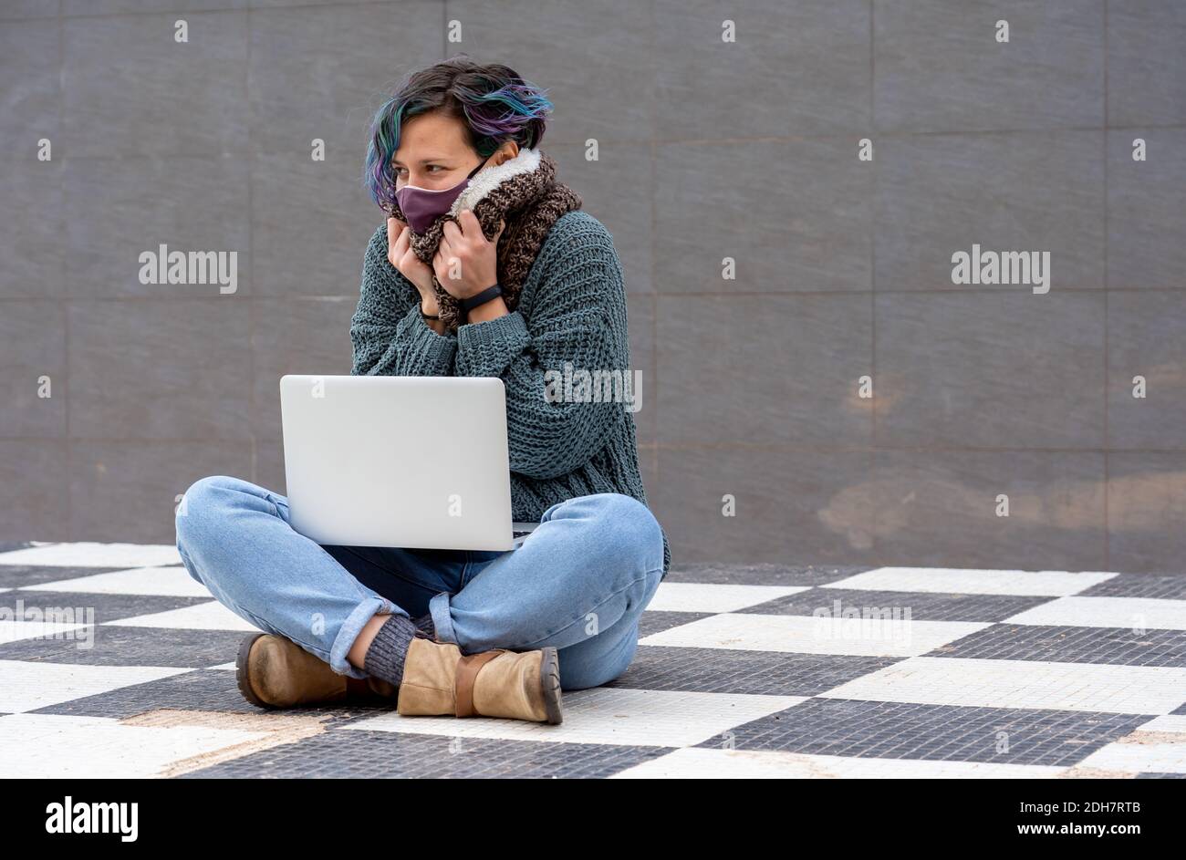 A stylish young lady sitting on a checkered floor in a park using her laptop wearing a sanitary mask Stock Photo