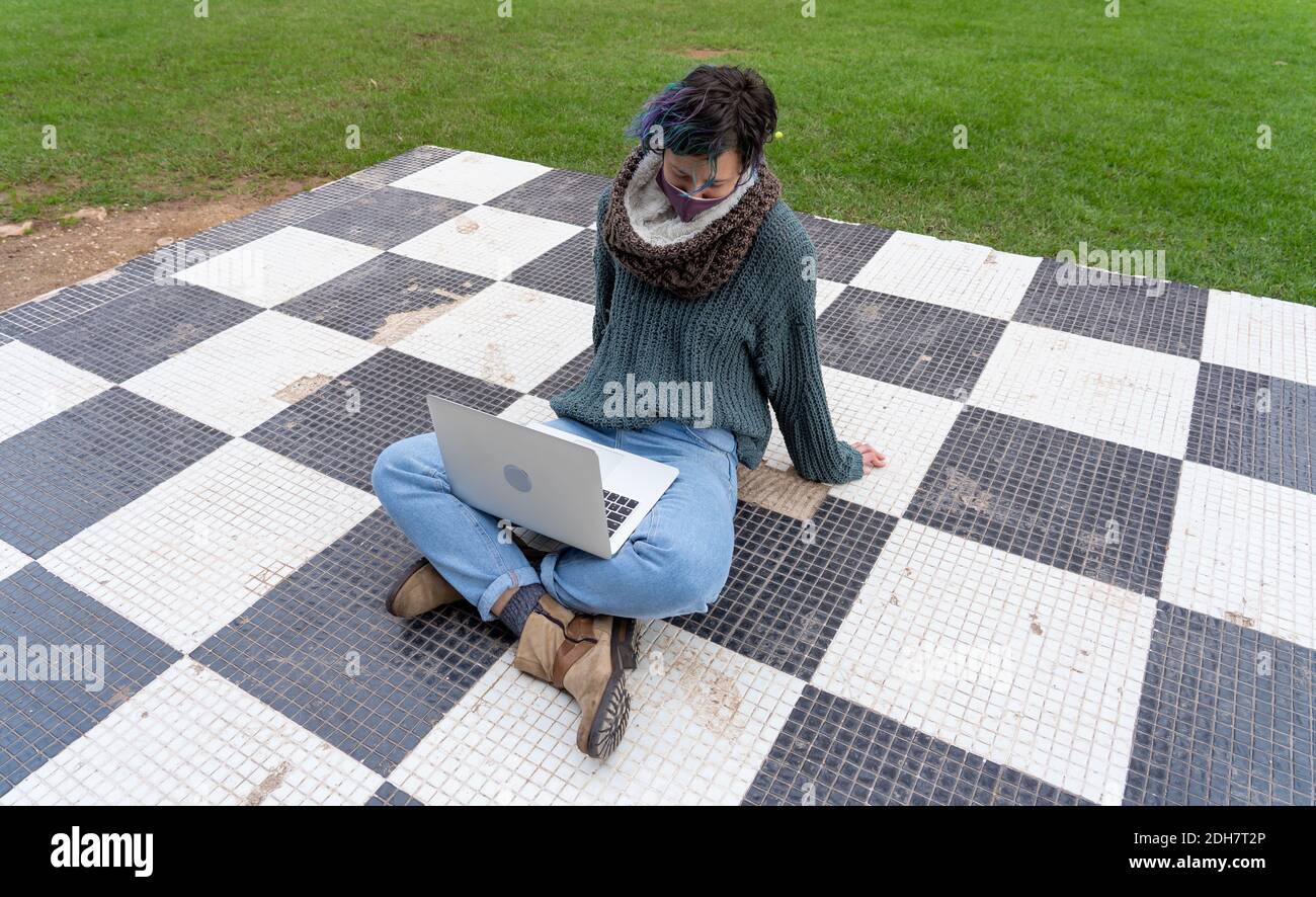 A stylish young lady sitting on a checkered floor in a park using her laptop wearing a sanitary mask Stock Photo