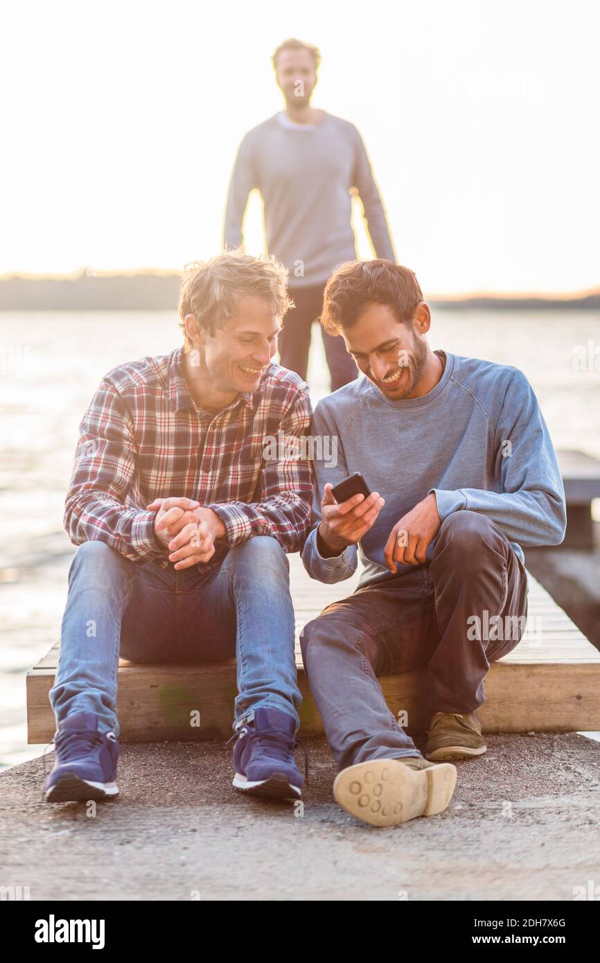 Happy male friends using mobile phone on pier with man walking in background Stock Photo
