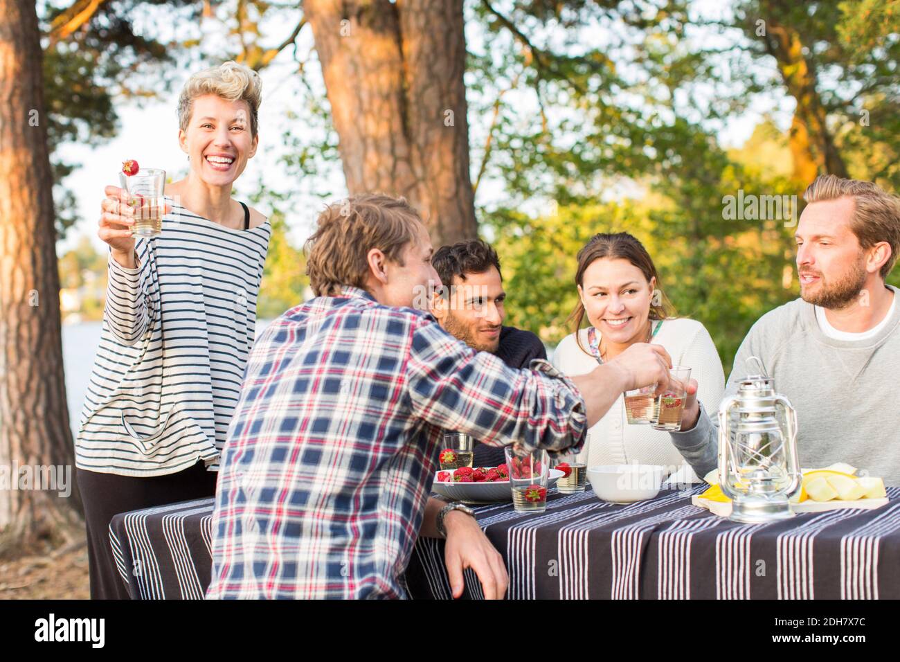 Portrait of happy woman holding beer glass while standing by friends having lunch at picnic table Stock Photo