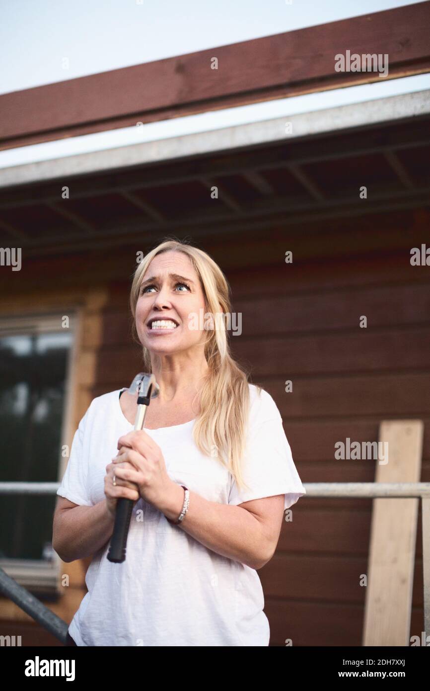 Angry woman clenching teeth while holding hammer outside house being renovated Stock Photo