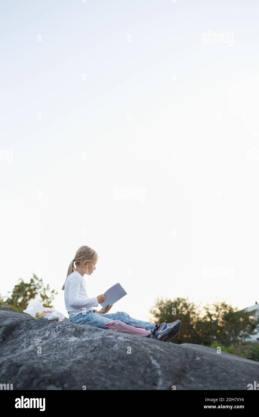 Full length side view of girl doing homework while sitting on rock against clear sky Stock Photo