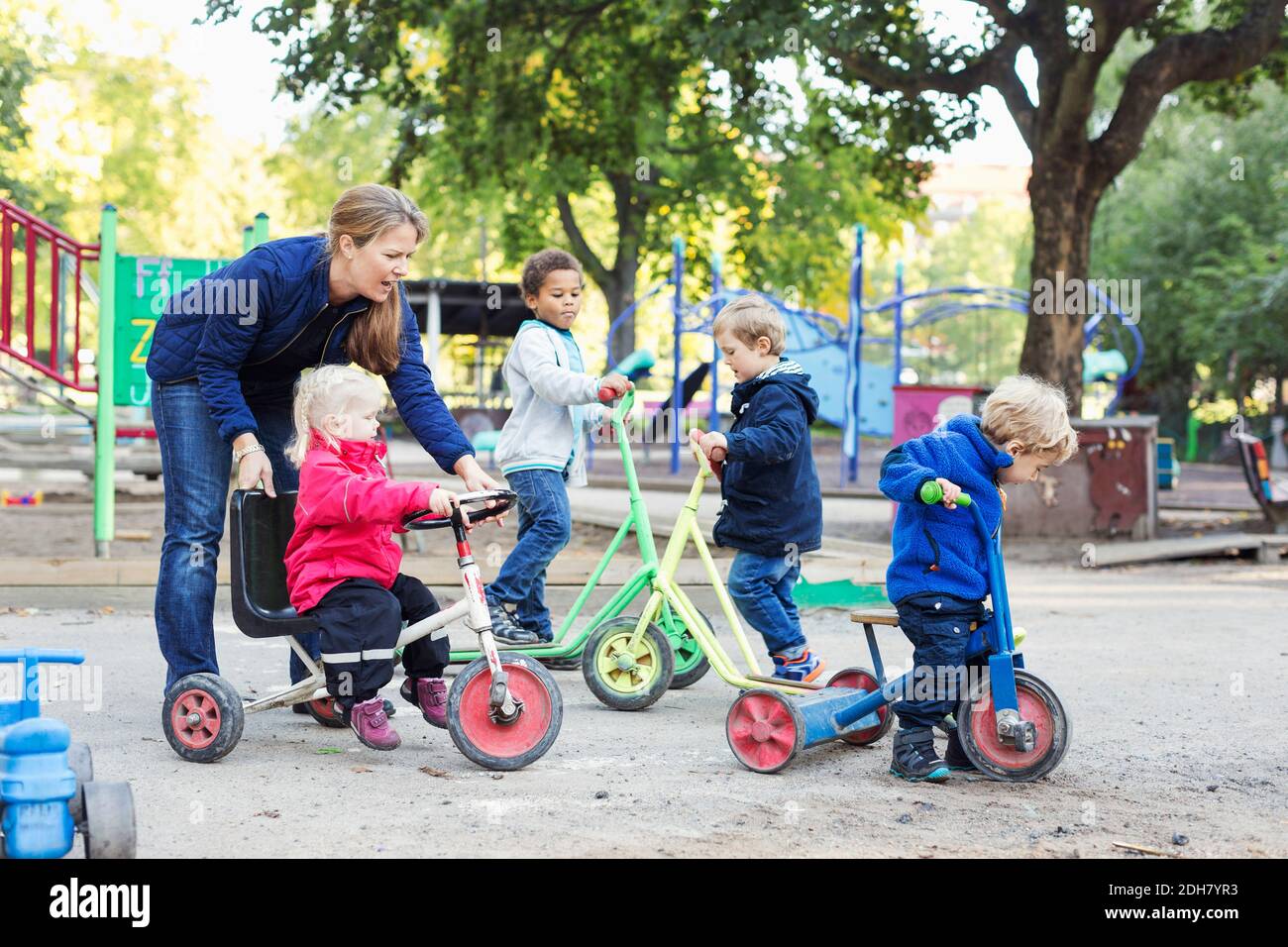 Female teacher assisting girl to ride tricycle on playground Stock Photo