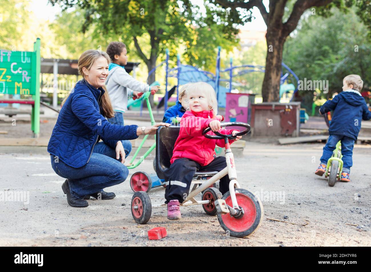 Happy female teacher assisting girl to ride tricycle on playground Stock Photo