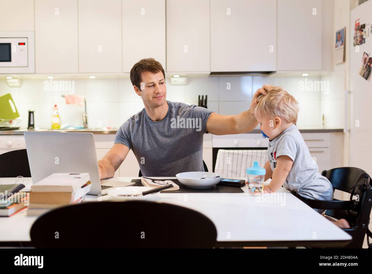 Father stroking baby boy while using laptop at dining table Stock Photo
