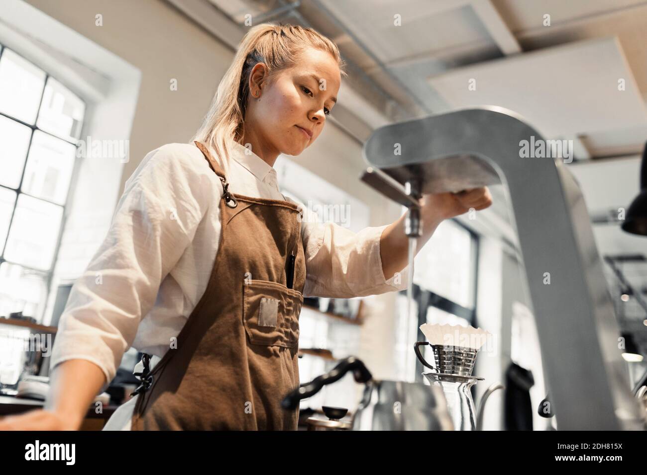 Young female barista pouring boiling water in coffee pot from machine
