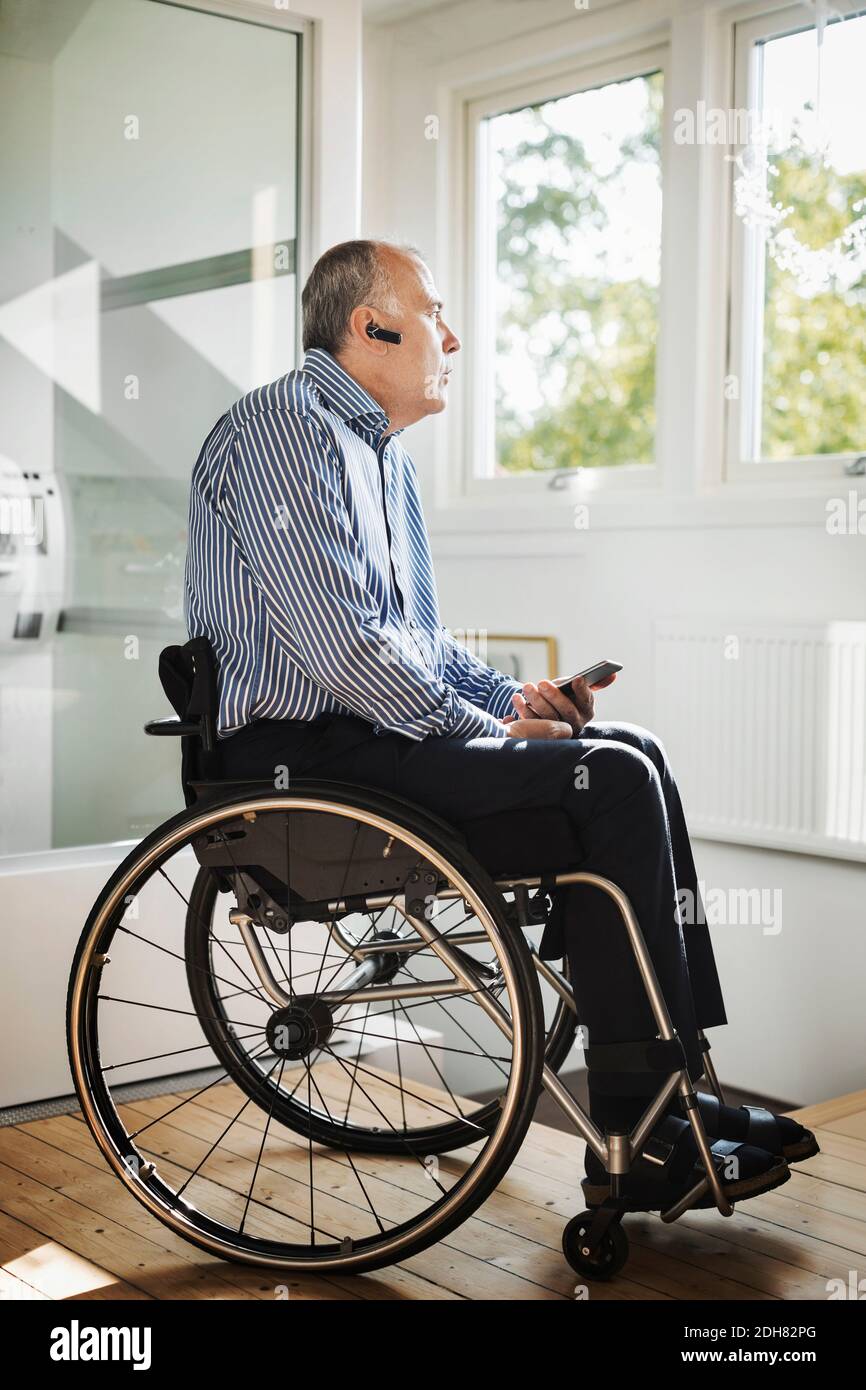 Mature man holding mobile phone while using Bluetooth in wheelchair at home Stock Photo