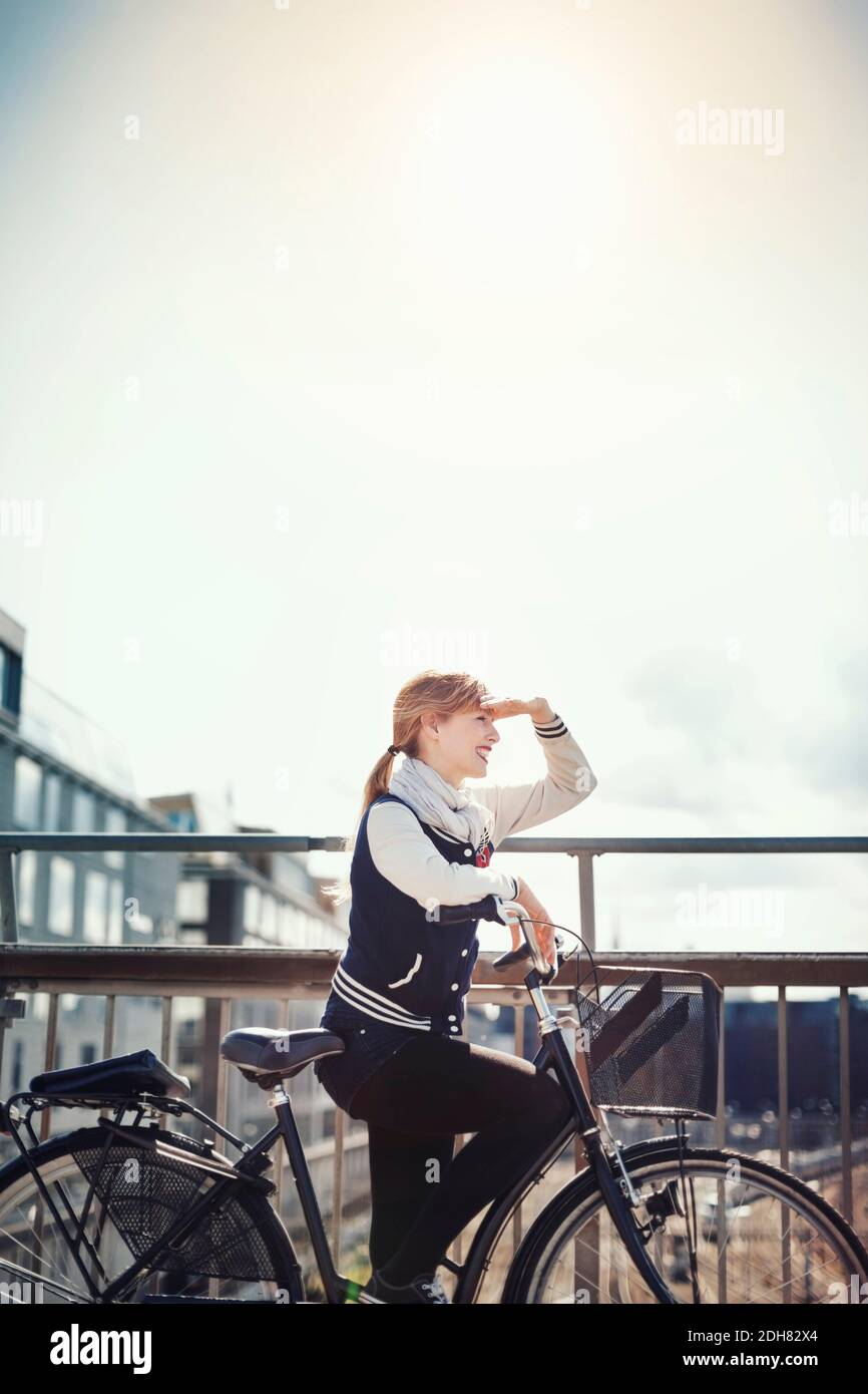 Happy businesswoman shielding eyes while standing with bicycle on bridge against sky Stock Photo