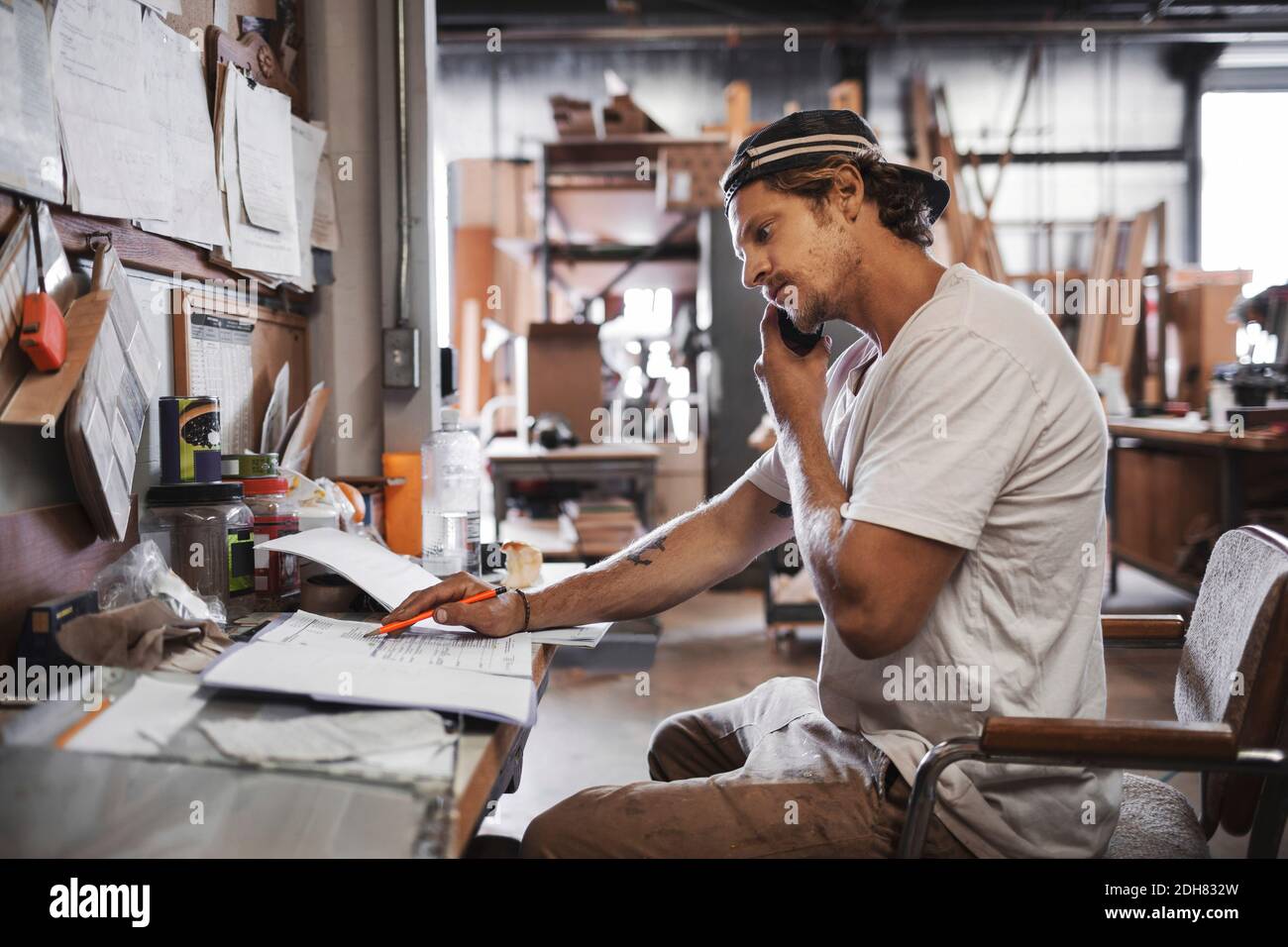 Side view of carpenter using mobile phone while reading document in workshop Stock Photo