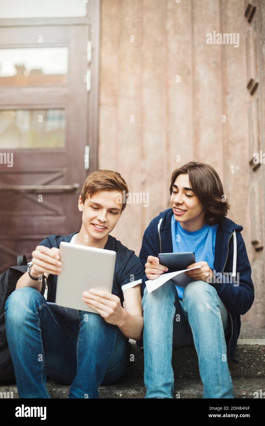 Happy male teenagers using digital tablet while sitting on steps outdoors Stock Photo