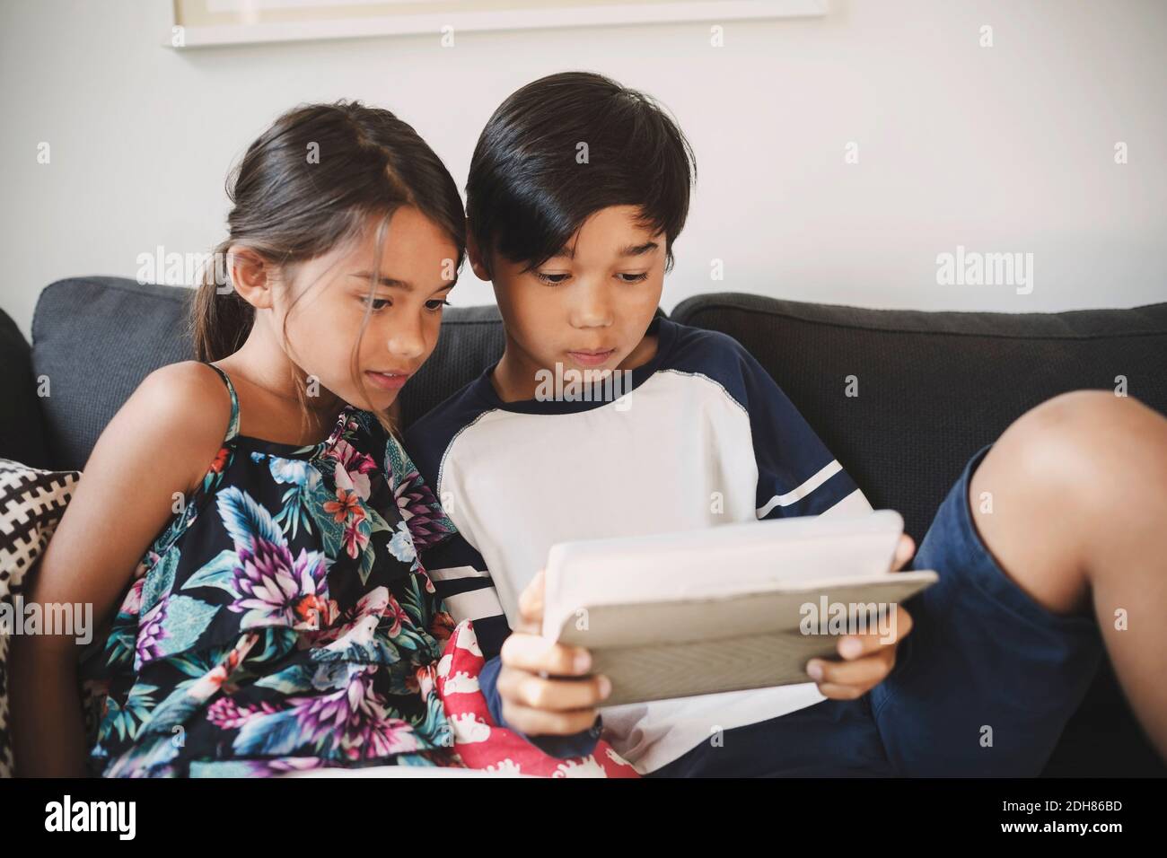 Brother and sister using digital tablet while sitting on sofa at home ...