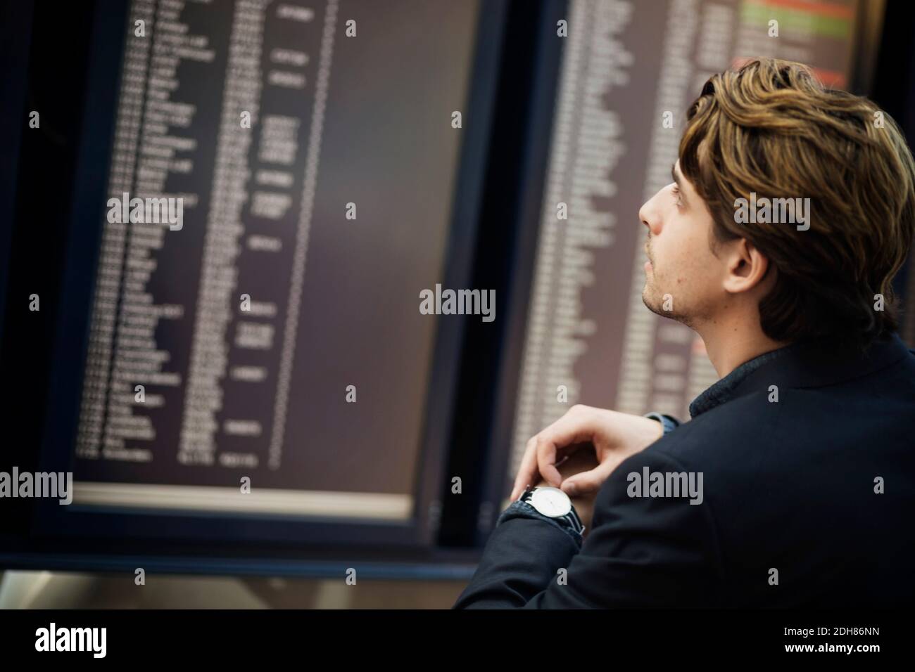 Businessman adjusting wrist watch while reading arrival departure board at airport Stock Photo