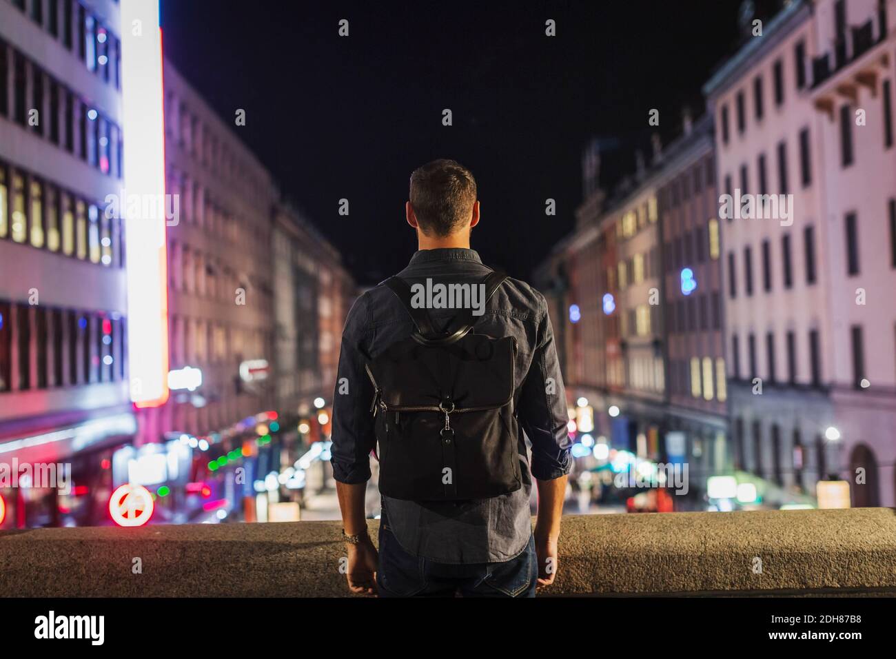 Rear view of man carrying backpack standing on bridge in city at night Stock Photo