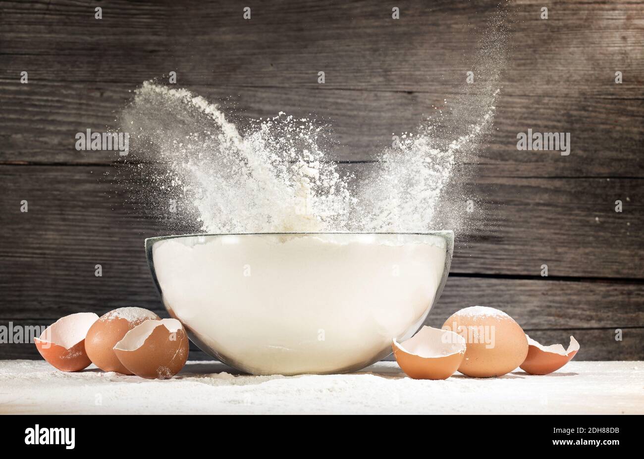 white flour splashing out of a glass bowl and eggshells on rustic background Stock Photo