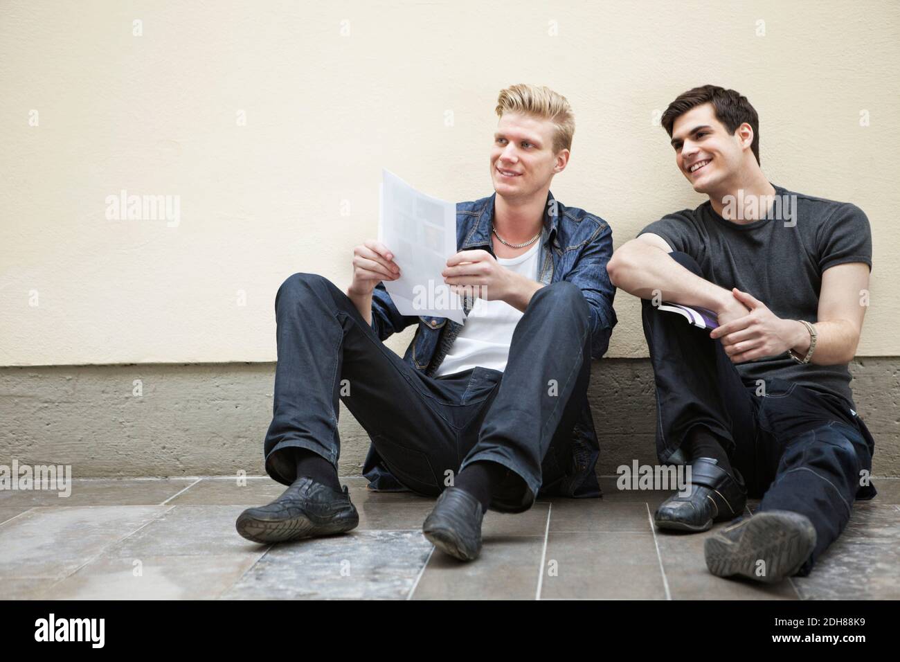 Full length of young male friends sitting down on floor looking away and smiling Stock Photo