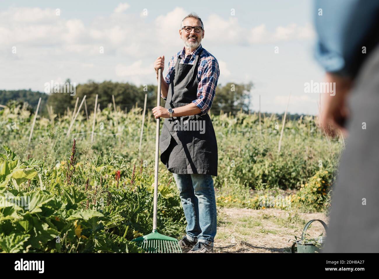 Mature farmer holding gardening fork and standing with coworker at farm ...